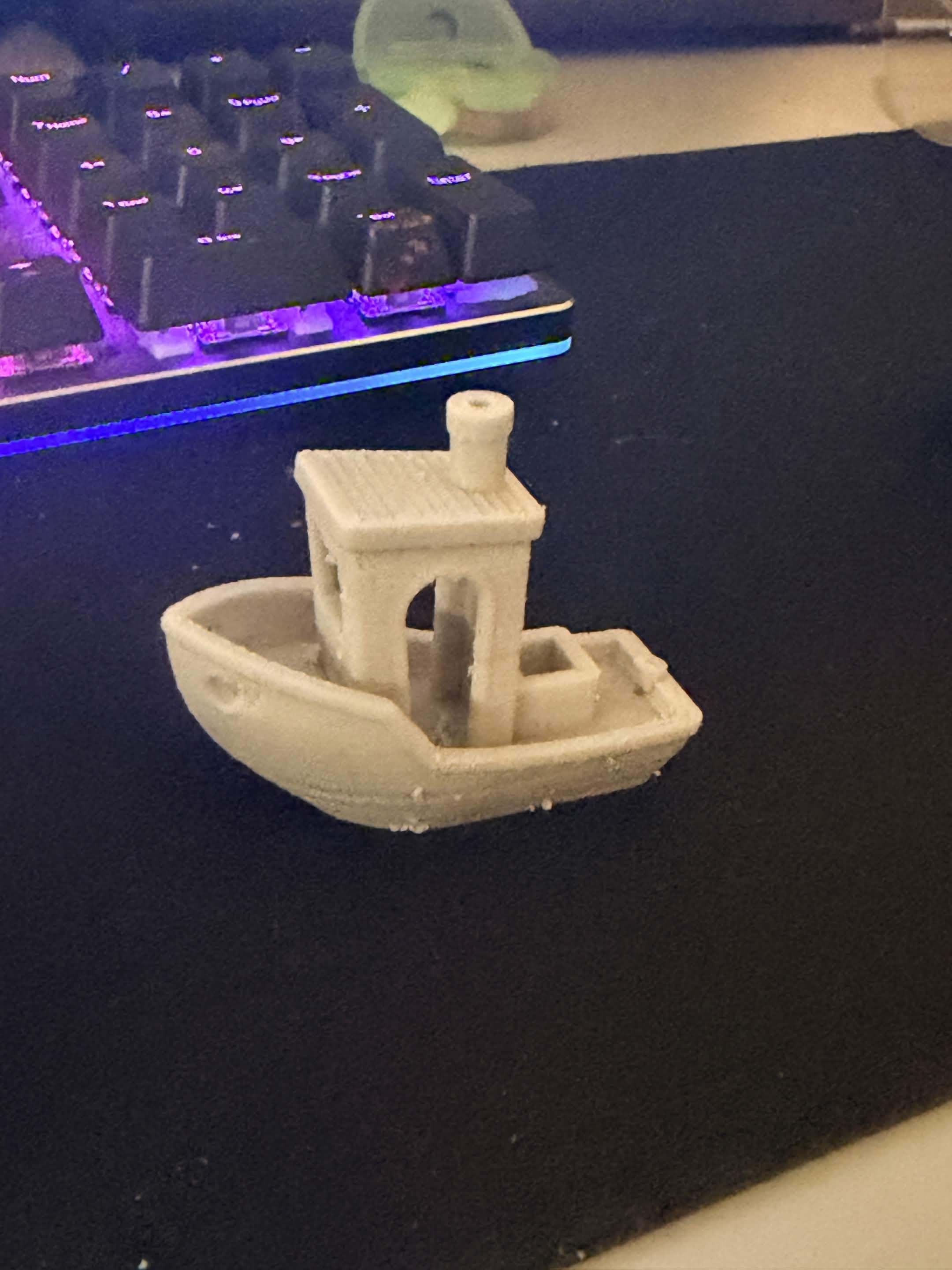 A 3DBenchy boat printed in borosilicate glass-filled filament, matte off-white, sitting on a desk mat.