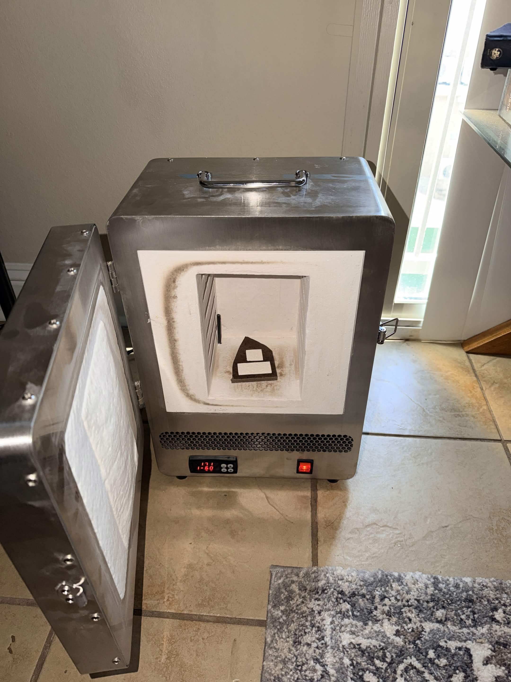 A small stainless bench kiln with its door open, showing a fired dark test article on white refractory board inside, controller reading temperature.